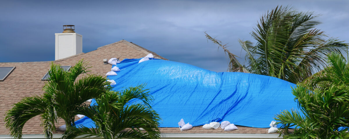 Storm damaged roof on house with a blue plastic tarp over hole in the shingles and rooftop.
